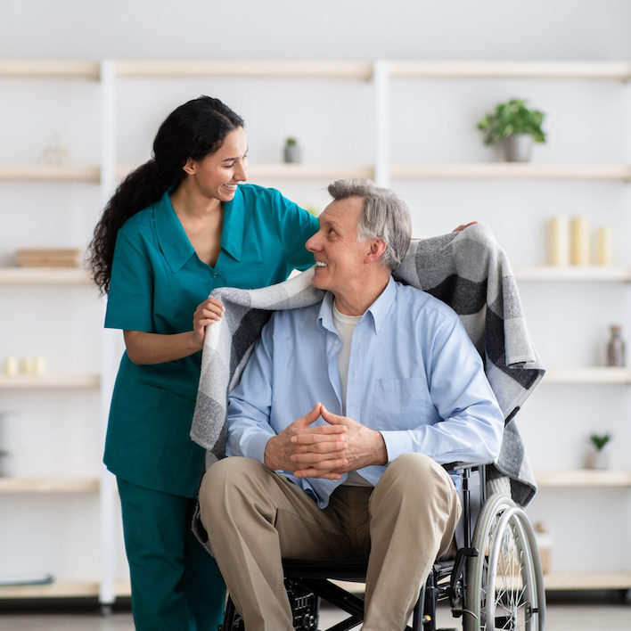 Young nurse covering disabled elderly man in wheelchair with warm plaid at retirement home. Millennial caregiver assisting handicapped senior patient, taking care of older male indoors